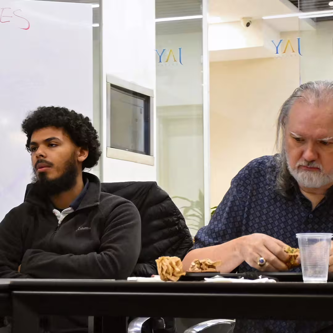 Davin Church (right) seated at a table during DYNA Spring 2025 in New York, focused on unwrapping food. He is wearing a patterned shirt. To his left, another attendee in a dark Columbia fleece sits with arms folded, listening attentively. Crumpled napkins and a plastic cup are visible on the table.