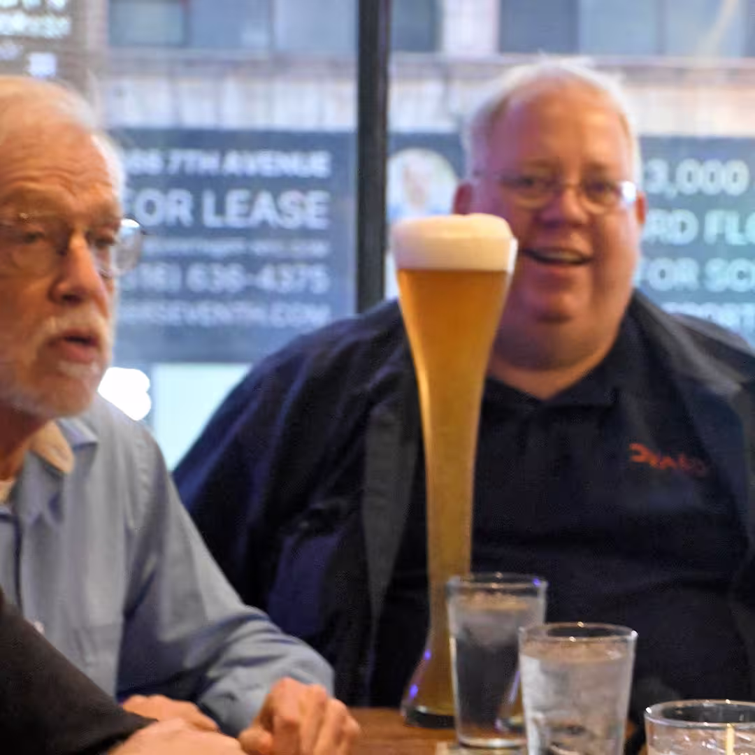 Brian Becker (right) enjoying a social gathering during DYNA Spring 2025 in New York. He is smiling and seated at a table with a tall glass of beer and water glasses in front of him. Another attendee is visible on the left, mid-conversation. A city street can be seen through the window behind them.