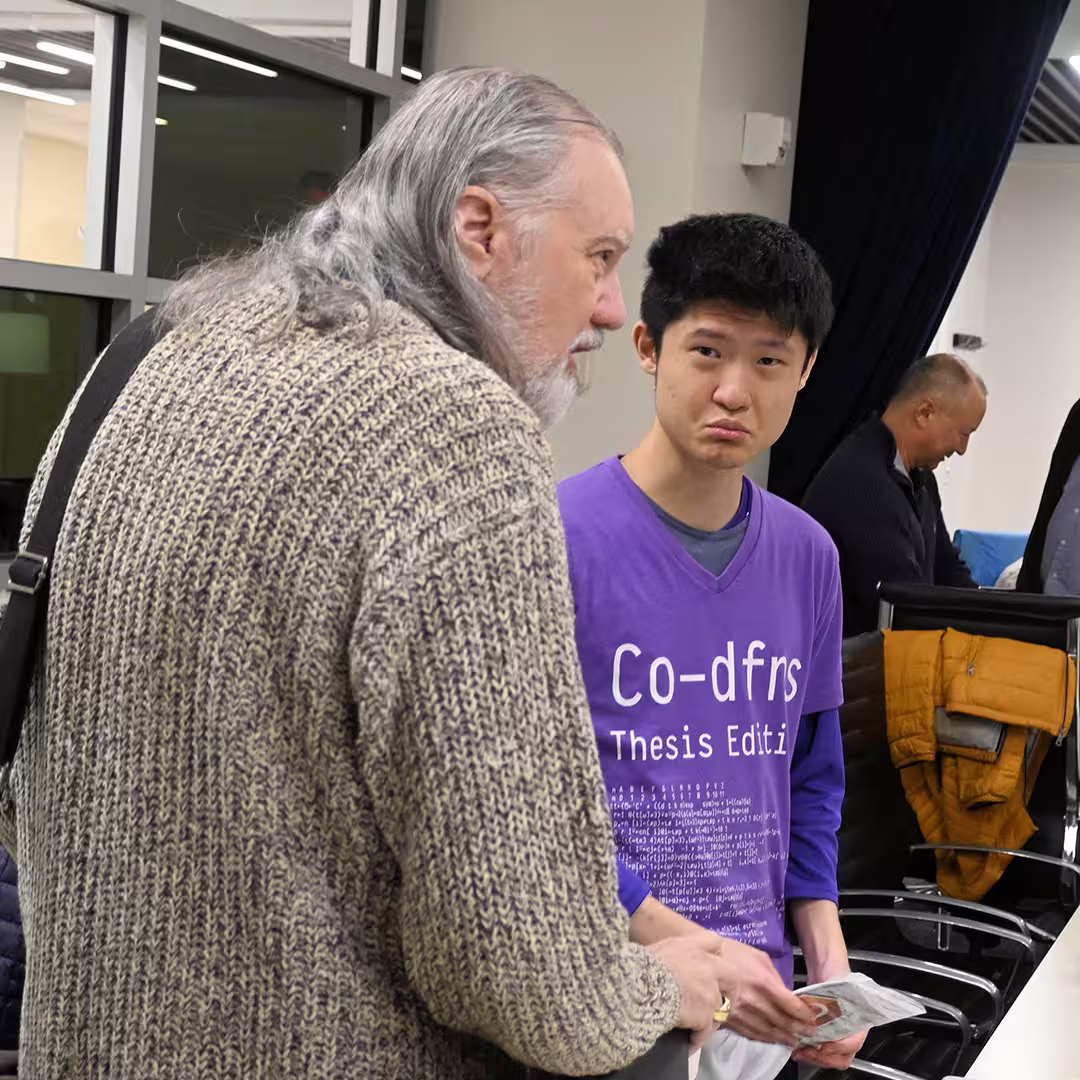 Davin Church and Max Sun at DYNA Spring 2025 in New York. Davin, wearing a chunky knit jumper and carrying a shoulder bag, is speaking with Max, who is wearing a purple "Co-dfns Thesis Edition" T-shirt and holding a paper. Other attendees can be seen in the background near a row of chairs.