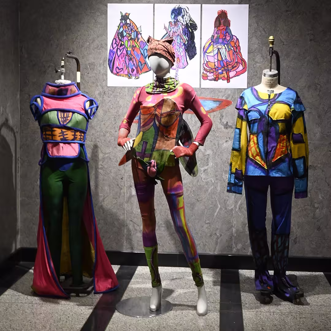 A display of three colourful, abstract costumes on mannequins at DYNA Spring 2025 in New York. Each outfit features vibrant, hand-painted patterns with a futuristic and whimsical design. Behind the mannequins are matching concept illustrations pinned to the wall, showcasing the artistic process behind the garments.