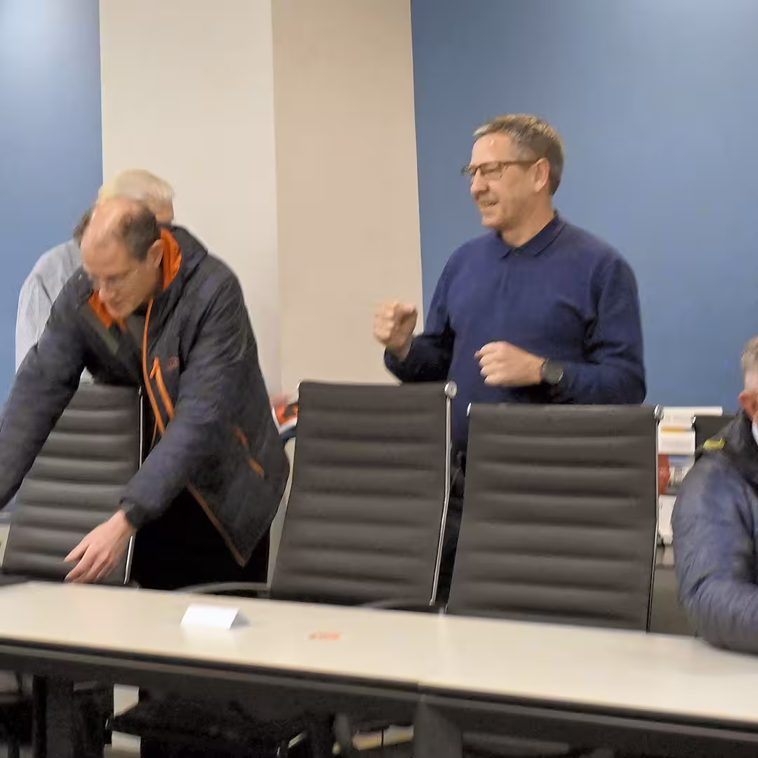 Morten Kromberg (centre) standing and gesturing mid-conversation during DYNA Spring 2025 in New York. He is wearing a dark blue top and glasses. Around him, other attendees are arriving or getting seated, with one person leaning over a chair in the foreground.