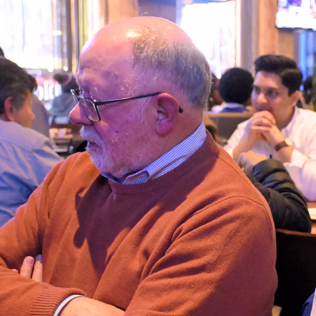 Jim Brown at a social gathering during DYNA Spring 2025 in New York. He is wearing glasses and a rust-coloured jumper, seated with arms crossed. In the background on the left is Mark Wolfson in a blue shirt, and on the right is Josh David in a white shirt, both seated and engaged in conversation