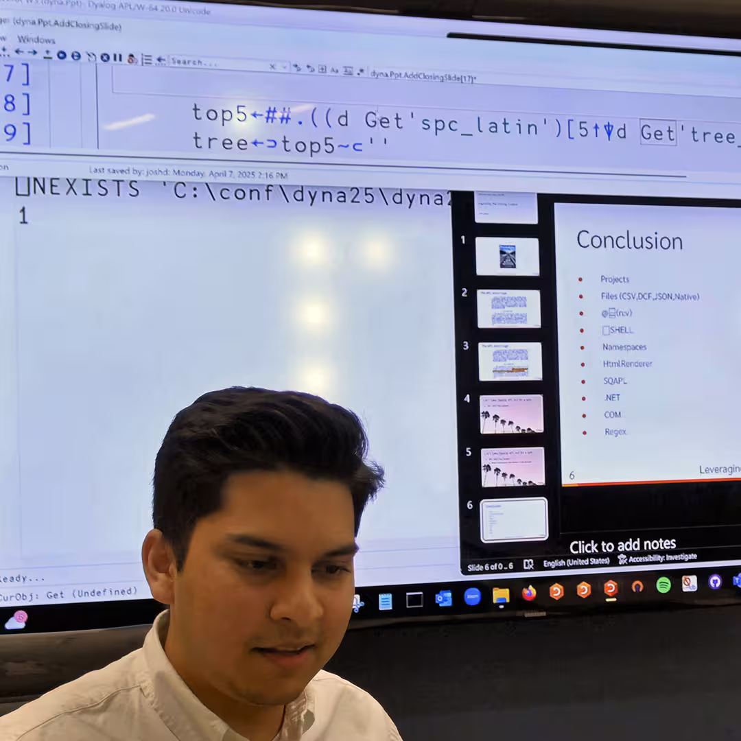 Josh David presenting at DYNA Spring 2025 in New York. He stands in front of a large screen displaying APL code and a slide titled “Conclusion,” listing various project components. Josh is wearing a white shirt and appears focused as he explains his material.