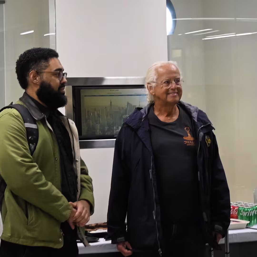 Lucas Santos and Chris Lincoln at DYNA Spring 2025 in New York. Lucas is wearing glasses, a green jacket, and a backpack, while Chris stands beside him in a dark jacket and Dyalog T-shirt, smiling. A table with snacks and drinks is visible in the background.